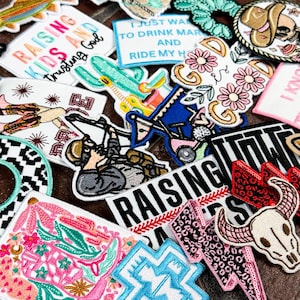 Könnte beinhalten: Eine Sammlung bunter gestickter Bügelbilder mit verschiedenen Designs, darunter eine Kuh, ein Kaktus, ein Stiefel, ein Schädel, ein Blitz, ein Kreuz und Text, der "Raising Kids and Trusting God" und "Raising Wild Ones" lautet.