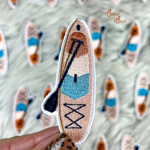 Könnte beinhalten: Gestickter Aufnäher mit einem Paddleboard-Design und einem Paddel. Der Aufnäher hat einen beigen Hintergrund mit blauen, orangefarbenen und weißen Akzenten. Das Paddel ist schwarz. Der Aufnäher wird von einer Hand mit einem Leopardenmuster-Nageldesign gehalten. Die Aufnäher sind auf einer weißen, flauschigen Oberfläche verstreut.