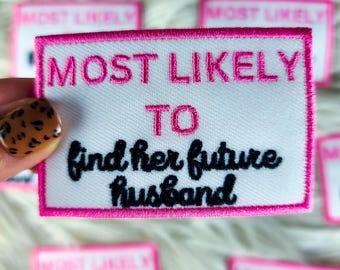Puede incluir: Parche rectangular blanco con borde bordado rosa y texto. El texto dice "MOST LIKELY TO find her future husband" en rosa y negro. Varios parches similares son visibles en el fondo.