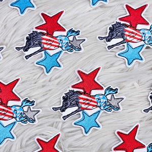 Può includere: Più patch da stirare che raffigurano un cavallo nero con strisce rosso, bianco e blu che salta sopra una stella rossa e una stella blu. I patch sono su una superficie bianca e soffice.