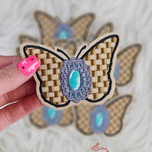 May include: A gold and brown checkered butterfly patch with a turquoise gem in the center. The butterfly has a black outline and is surrounded by other butterfly patches.