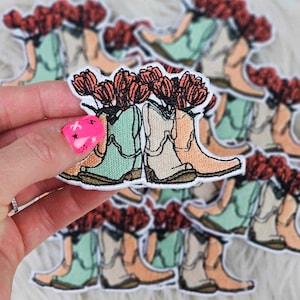 May include: A set of two embroidered iron-on patches featuring a pair of cowboy boots with a bouquet of red flowers on top. The boots are in shades of green, beige, and brown.