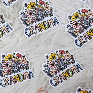 Peut inclure: Un ensemble de patchs brodés colorés à repasser avec le mot "Grandma" et un motif floral. Les patchs sont sur une surface blanche et duveteuse.