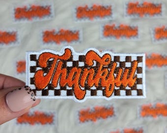 Peut inclure: Écusson brodé avec le mot "Thankful" en écriture orange sur un fond à carreaux marron et blanc. L'écusson est rectangulaire avec une bordure blanche. Plusieurs écussons similaires sont en arrière-plan.