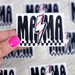 以下が含まれることがあります： 白地に黒文字で「MAMA」と書いてあるアイロン接着パッチ。中央には赤色のステッチで縫われた白い稲妻と野球のデザインが施されています。パッチは黒と白の市松模様の縁取りがされています。