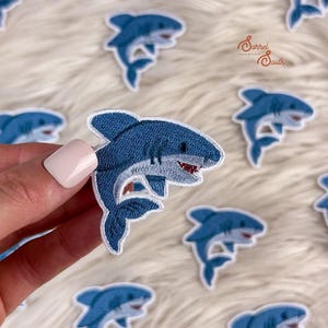 May include: Embroidered shark patches in shades of blue and white. The patches feature a detailed design of a shark with a white belly and red mouth. The patches are on a white, fluffy surface.