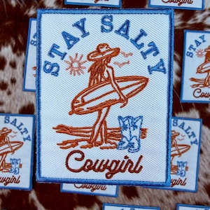 May include: Embroidered patch with a white background, blue border, and the words "Stay Salty" in blue. The patch features a cowgirl with a surfboard, a sun, and the word "Cowgirl" in brown.