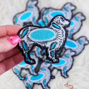 May include: Embroidered dog patch with a black outline, white body, and turquoise accents. The patch features a detailed design with a turquoise oval in the center and turquoise shapes along the body. Several patches are visible.