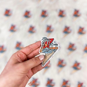 May include: A colorful embroidered patch featuring a tropical beach scene with palm trees, waves, and a sunset. The patch is shaped like a lightning bolt and has a white border. The background is a soft, white surface.