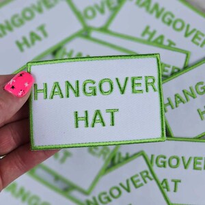 May include: White rectangular patch with the words "HANGOVER HAT" embroidered in green. The patch has a green border. Multiple patches are visible, some out of focus.