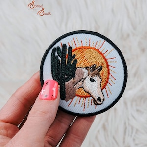 May include: A circular embroidered patch featuring a brown and white horse head positioned in front of a dark green cactus and a bright orange sun with radiating rays. The patch has a thick black border and the text Sorrel South in the upper left.