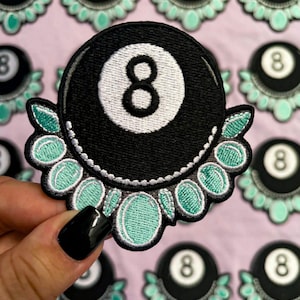 May include: A circular embroidered patch featuring a black eight ball with a white center circle and the number eight in black. The bottom half of the patch is decorated with a fan-shaped arrangement of light green oval shapes outlined in white thread.