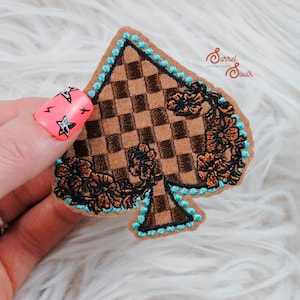 Może przedstawiać: Naszywka z brązowej i czarnej kraty w kształcie pika z turkusową lamówką. Pik ozdobiony jest brązowymi haftowanymi kwiatami.
