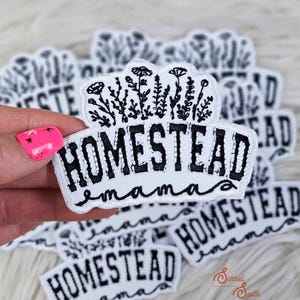 May include: White embroidered patch with the words "HOMESTEAD mama" in black script. The patch features a floral design at the top. Several similar patches are visible in the background.