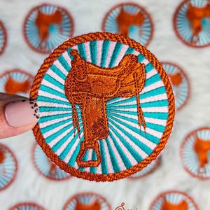 May include: Embroidered patch featuring a brown saddle design. The patch has a turquoise and pink striped background with a brown rope border. The design is circular and suitable for apparel or accessories.