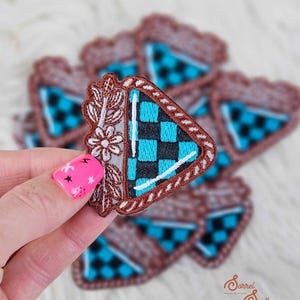 Pode incluir: Um remendo bordado marrom e branco com um design de triângulo xadrez azul e preto. O remendo tem um design floral no lado esquerdo e uma borda branca ao redor do triângulo.