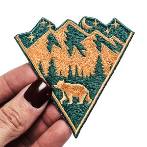 May include: A green and gold embroidered patch with a mountain range, trees, a bear, a crescent moon, and stars.