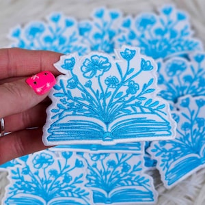 Op de afbeelding: Een stapel witte strijkbare patches met blauwe borduursels van een open boek met bloemen die eruit groeien.