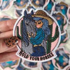 May include: Embroidered patch with a cowgirl in a brown hat and blue hair embracing a grey horse, set against a pink border and green cacti. The text "HOLD YOUR HORSES" is at the bottom.