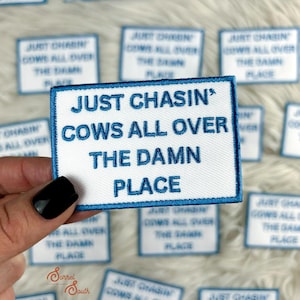 Puede incluir: Parche bordado blanco con texto azul: "JUST CHASIN' COWS ALL OVER THE DAMN PLACE." El parche tiene un borde azul. Varios parches similares son visibles en el fondo.
