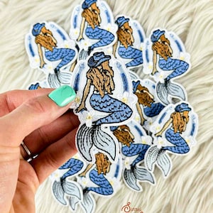 May include: Embroidered patches featuring a mermaid with long auburn hair, wearing a blue cowboy hat and a blue scaled tail. The patches have a white border and are decorated with white flowers. The patches are held in a hand.