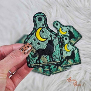 以下が含まれることがあります： 背景に星が描かれた、月に向かって吠えるオオカミがデザインされた、緑色の刺繍パッチ4枚セット。パッチは牛のタグの形をしており、ステッチで縁取られています。