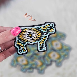 May include: A turquoise, yellow, and white embroidered iron-on patch of a cow. The patch is on a white background.