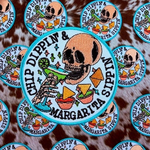 Może przedstawiać: Haftowana naszywka z jasnoniebieską obwódką przedstawiająca czaszkę pijącą margaritę, z napisem "Chip Dippin' & Margarita Sippin'". Projekt zawiera chipsy, salsę i szklankę do margarity. Naszywki znajdują się na brązowym i białym tle ze skóry krowy.