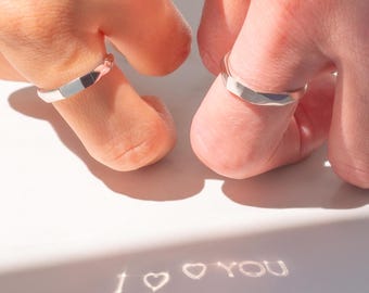 May include: Two silver rings on fingers, with the reflection of "I love you" on a white surface.