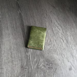 May include: Olive green leather cardholder with a minimalist design. Features a diagonal pocket and red stitching along the bottom edge. A small emblem is visible on the lower right corner. The cardholder is rectangular and appears to be made of textured leather.