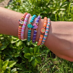 May include: Five colorful beaded bracelets with different fruit charms. The bracelets are stacked on a person's wrist. The charms include an apple, a flower, a strawberry, and an orange slice.