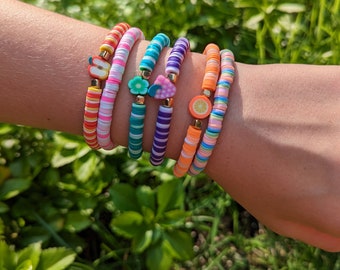May include: Five colorful beaded bracelets with different fruit charms. The bracelets are stacked on a person's wrist. The charms include an apple, a flower, a strawberry, and an orange slice.