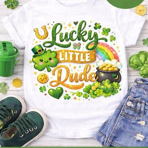 May include: White t-shirt with a St. Patrick's Day design. The shirt features the text "Lucky Little Dude" in green and gold, along with a rainbow, shamrocks, a pot of gold, and a leprechaun. The image also includes green shoes and denim shorts.