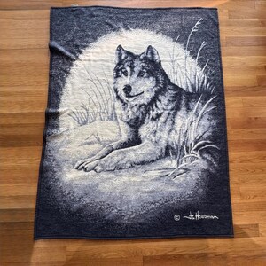 May include: A tapestry blanket with a detailed image of a wolf resting in a grassy area. The blanket has a dark blue border and a light background with the wolf in shades of grey and white. The artist's signature is in the bottom right corner.