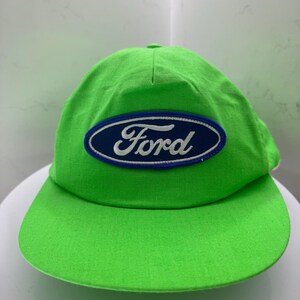 May include: A vivid green baseball cap with a curved peak. The cap displays the Ford logo in a navy blue oval with a white border. The cap is made of a soft material and has a classic, casual design.