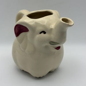 Peut inclure: Pichet en céramique crème en forme d'éléphant, avec un bec et une anse. L'éléphant a des accents rouge foncé sur les oreilles et la bouche, avec des yeux noirs. Objet décoratif fantaisiste.