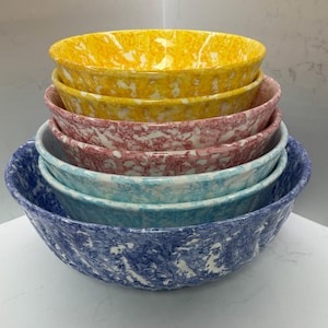 Peut inclure: Un ensemble de quatre bols mélangeurs emboîtables présentant un motif marbré et tacheté. Les bols sont empilés du plus petit au plus grand, affichant des couleurs vives dont le jaune, le rose, le bleu clair et le bleu foncé, avec des touches blanches.