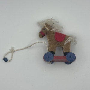 Puede incluir: Un pequeño caballo de juguete para tirar, con cuerpo marrón, crin y cola blancas y una silla de montar roja. El caballo está montado sobre una base de madera roja con ruedas azules. Se adjunta una cuerda blanca para tirar.