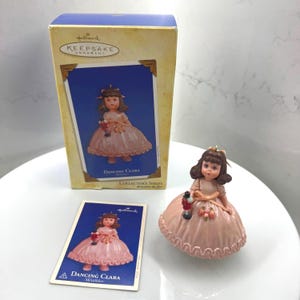 Könnte beinhalten: Ein Hallmark Keepsake Ornament einer Puppe namens "Dancing Clara" in einem rosa Kleid, die einen Nussknacker hält. Das Ornament wird mit der Originalverpackung und einer Sammlerkarte präsentiert. Die Box hat das Hallmark Keepsake Ornament Logo.