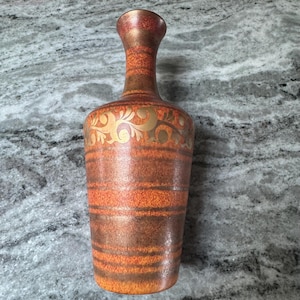Könnte beinhalten: Eine hohe Keramikvase mit schmalem Hals und breitem Boden. Die Vase hat horizontale Bänder in Orange, Braun und Gold. Goldene florale Designs verzieren den oberen Teil. Die Vase hat eine glänzende Oberfläche.