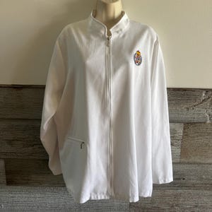 May include: A white long-sleeve zip-up jacket featuring a mock neck collar and a small embroidered crest on the left chest. The crest displays a gold crown above a shield surrounded by a laurel wreath. It includes a single zippered pocket.