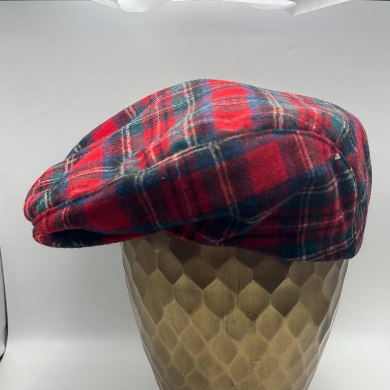 Op de afbeelding: Een platte pet met een veelkleurig ruitpatroon met opvallende rode, donkergroene en marineblauwe lijnen. De pet heeft een afgeronde, gestructureerde kroon en een korte, stijve klep aan de voorzijde, met een zachte textuur.