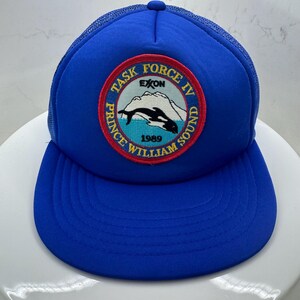 May include: A blue trucker hat with a curved brim and mesh back. The front features a circular patch with the text "TASK FORCE IV PRINCE WILLIAM SOUND" and a graphic of a whale and mountains. The year "1989" is also displayed.