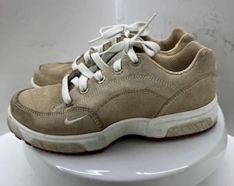 May include: A pair of tan suede athletic shoes with white laces and a white sole. The shoes have a small Nike swoosh logo on the side. The soles have a red accent.