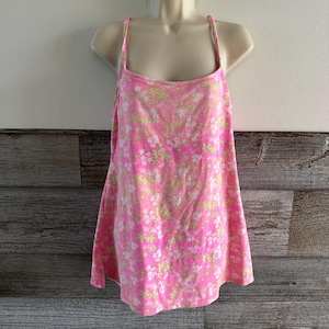K&ouml;nnte beinhalten: Ein rosa Camisole mit einem Blumenmuster in Wei&szlig; und Hellgr&uuml;n. Das Oberteil hat d&uuml;nne Spaghettitr&auml;ger und einen quadratischen Ausschnitt. Der Stoff scheint weich und leicht zu sein, geeignet f&uuml;r warmes Wetter.