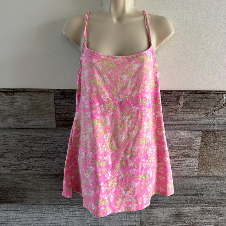 K&ouml;nnte beinhalten: Ein rosa Camisole mit einem Blumenmuster in Wei&szlig; und Hellgr&uuml;n. Das Oberteil hat d&uuml;nne Spaghettitr&auml;ger und einen quadratischen Ausschnitt. Der Stoff scheint weich und leicht zu sein, geeignet f&uuml;r warmes Wetter.