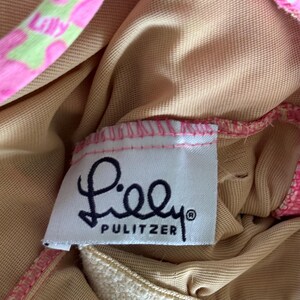 K&ouml;nnte beinhalten: Nahaufnahme eines wei&szlig;en Etiketts mit dem Markennamen "Lilly Pulitzer" in schwarzer Schrift. Das Etikett ist auf einen rosa gemusterten Stoff gen&auml;ht, mit einem beigefarbenen Futter. Der Stoff hat ein rosa-gr&uuml;nes Blumenmuster.