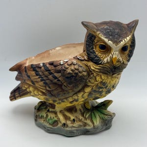 May include: A ceramic owl-shaped planter featuring detailed brown, black, and yellow plumage. The owl has large dark eyes, a yellow beak, and is perched on a textured base with green leaf accents. The interior of the planter is hollow and beige.