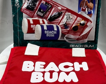 Pode incluir: Uma bolsa de praia vermelha com alças brancas e as palavras "BEACH BUM" em letras brancas grandes. A bolsa é combinada com uma sacola transparente contendo óculos de sol, uma garrafa preta e outros itens. Duas toalhas de praia enroladas, uma vermelha e outra azul, também estão incluídas.