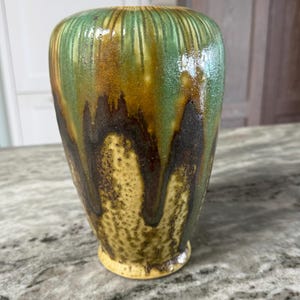 May include: A handcrafted ceramic vase with a unique, textured glaze. The vase features a blend of colors, including green, brown, and yellow, with a dripped effect. The vase has a rounded base and a slightly flared top, perfect for displaying flowers.